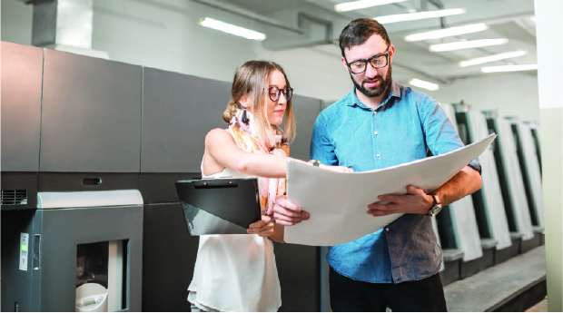 2 individuals studying a print with printing machine behind them 